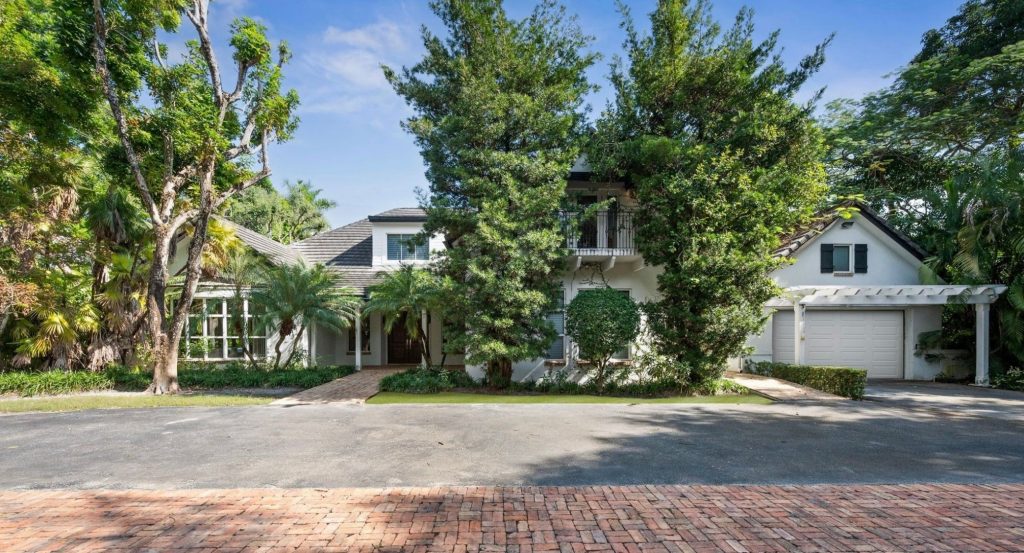 White two-story luxury home with dark tile roof, wraparound porch, attached garage with pergola, and lush tropical landscaping including mature trees and palm trees, viewed from a brick-paved street.