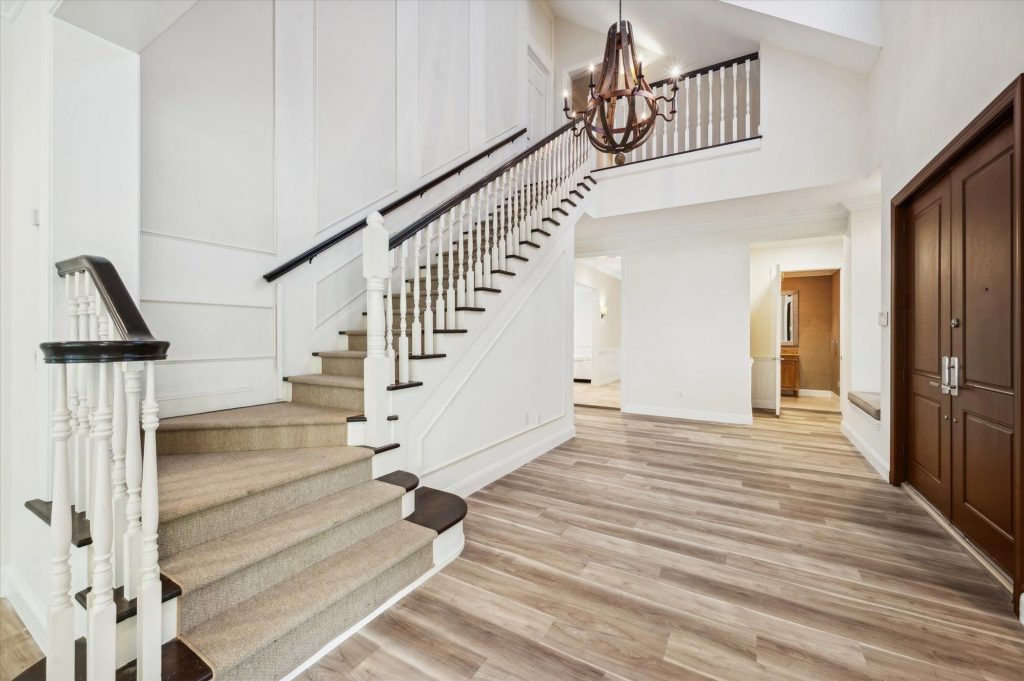 Grand two-story foyer featuring a carpeted staircase with white balusters and dark wood handrail, light hardwood floors, wainscoting wall paneling, a rustic wood chandelier, and a solid dark wood front door.