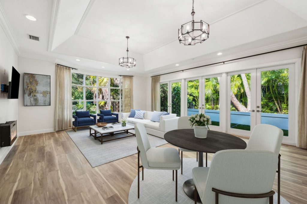 Bright open-plan living and dining area with light hardwood floors, tray ceiling, two geometric metal chandeliers, a white sectional sofa with blue accent chairs, dark oval dining table, and floor-to-ceiling French doors overlooking tropical greenery.