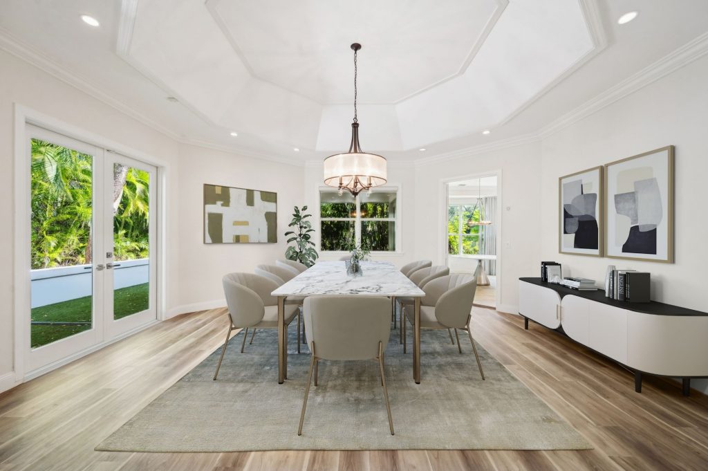 Elegant formal dining room with an octagonal coffered ceiling, marble-top rectangular dining table surrounded by eight upholstered chairs, a drum pendant light, abstract wall art, and French doors opening to a landscaped yard.