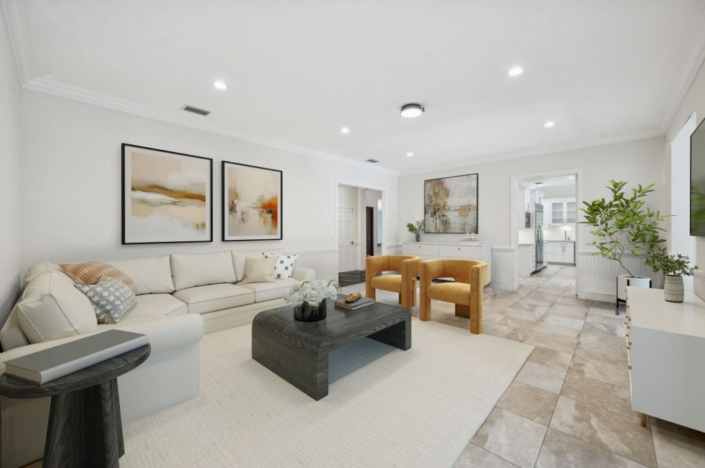 Spacious family room with large-format neutral tile flooring, cream sectional sofa, dark wood coffee table, mustard velvet accent chairs, warm abstract artwork, crown molding, and an open view into a white kitchen.