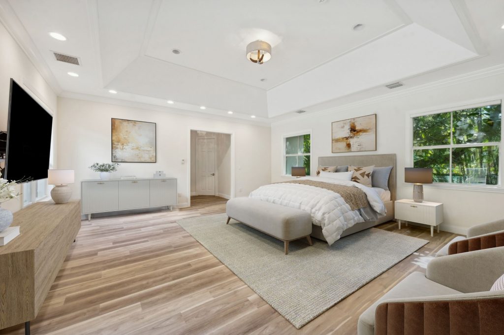 Expansive primary bedroom with tray ceiling, light hardwood floors, upholstered king bed, large wall-mounted TV, neutral abstract artwork, a sitting area with velvet chairs, and oversized windows with tropical garden views.