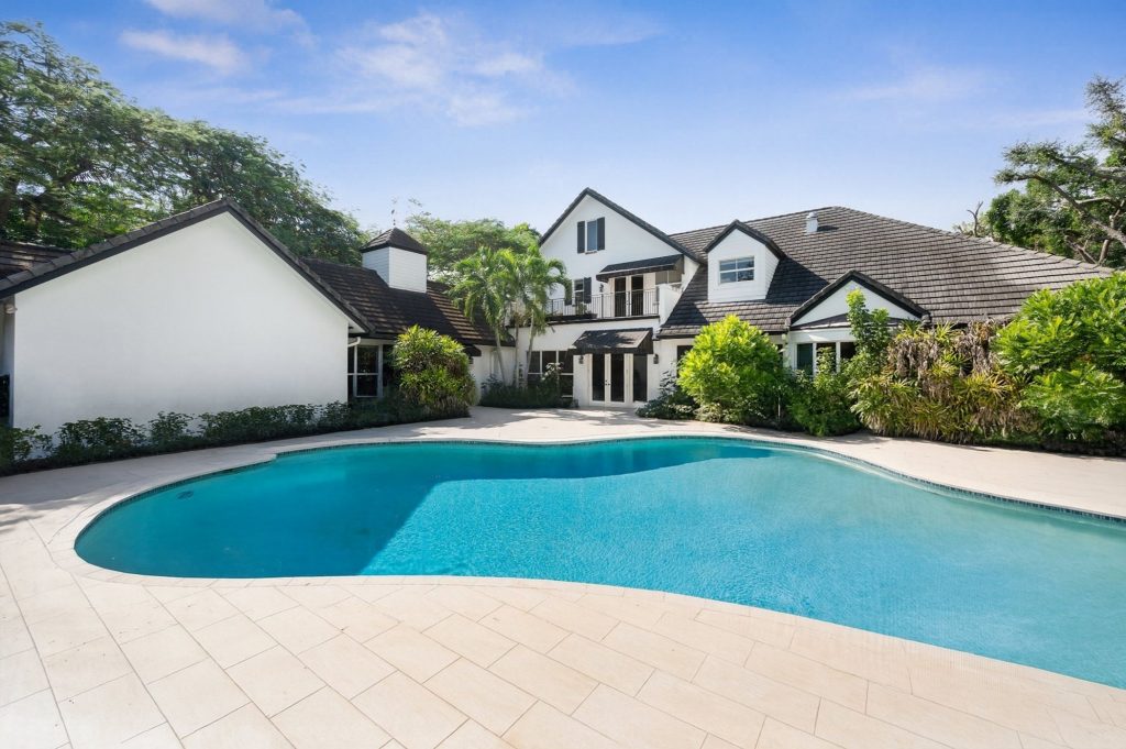  Rear exterior of a white two-story home with dark tile roof and upper balcony, surrounded by lush tropical landscaping, featuring a large freeform swimming pool with a travertine pool deck.